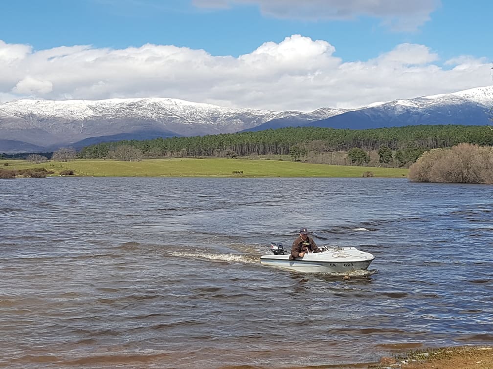 Persona en un pequeño bote motorizado en un lago, con montañas nevadas y un campo verde al fondo.