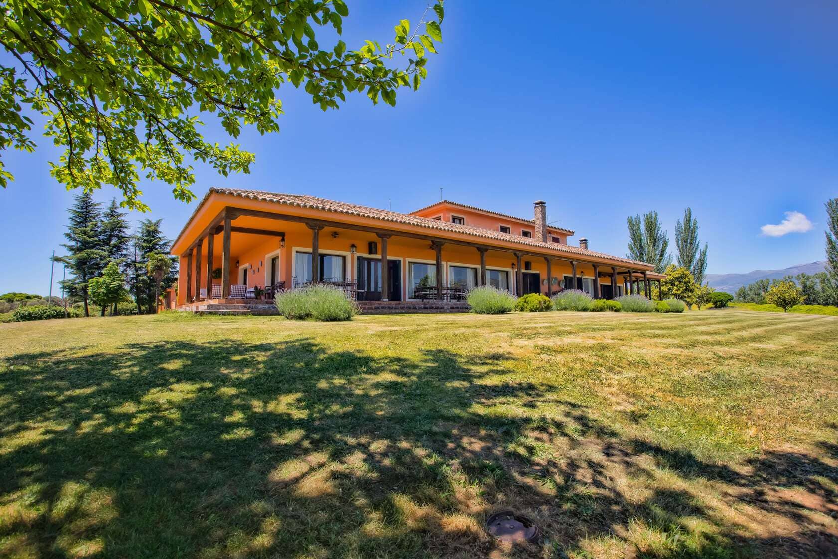 Vista de una amplia casa rural con paredes de color naranja, rodeada de un jardín verde y árboles, bajo un cielo despejado.