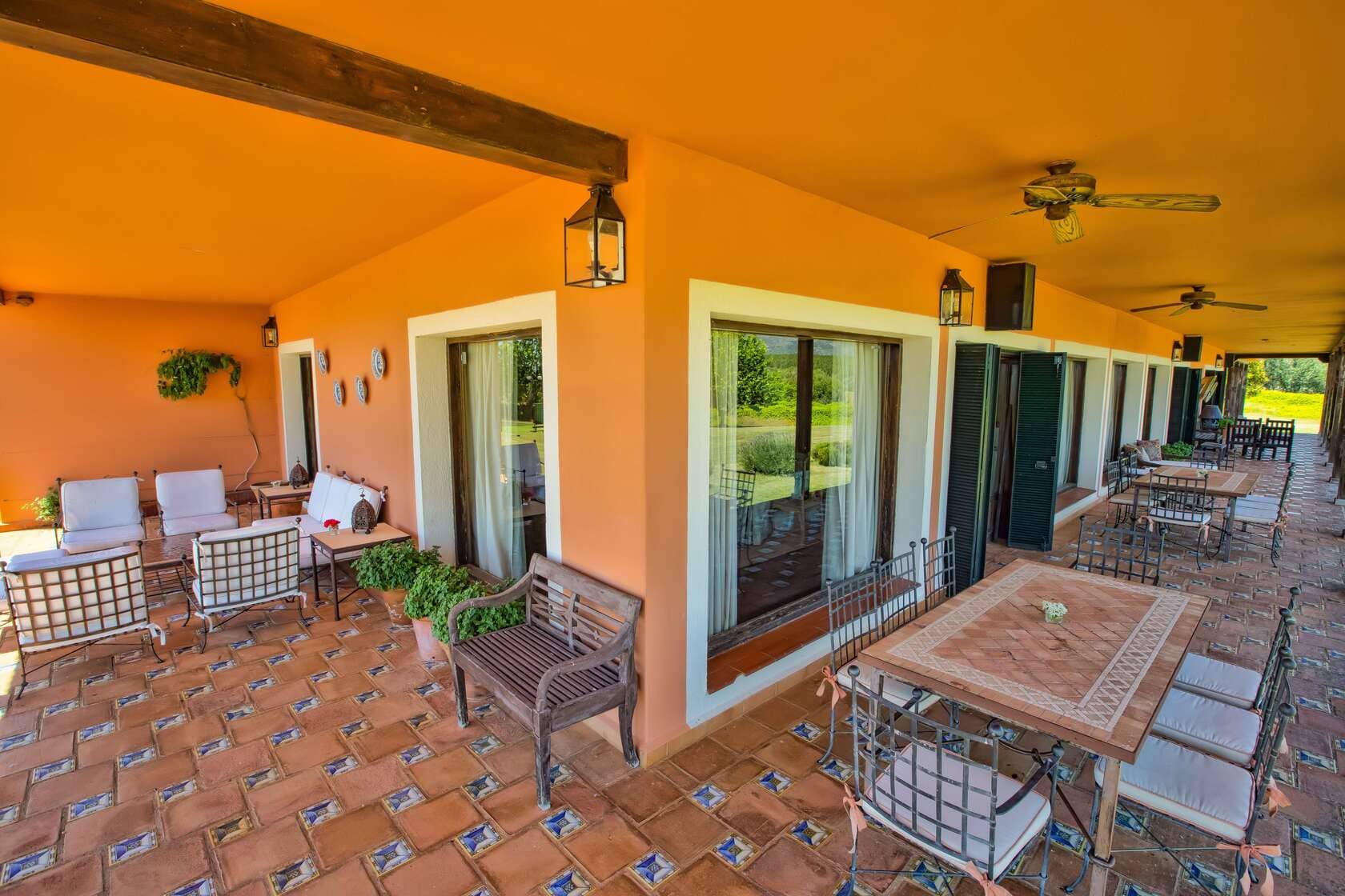 Una terraza exterior con muebles de hierro forjado y cojines blancos, decorada con plantas y cerámicas, en una casa de paredes color naranja.