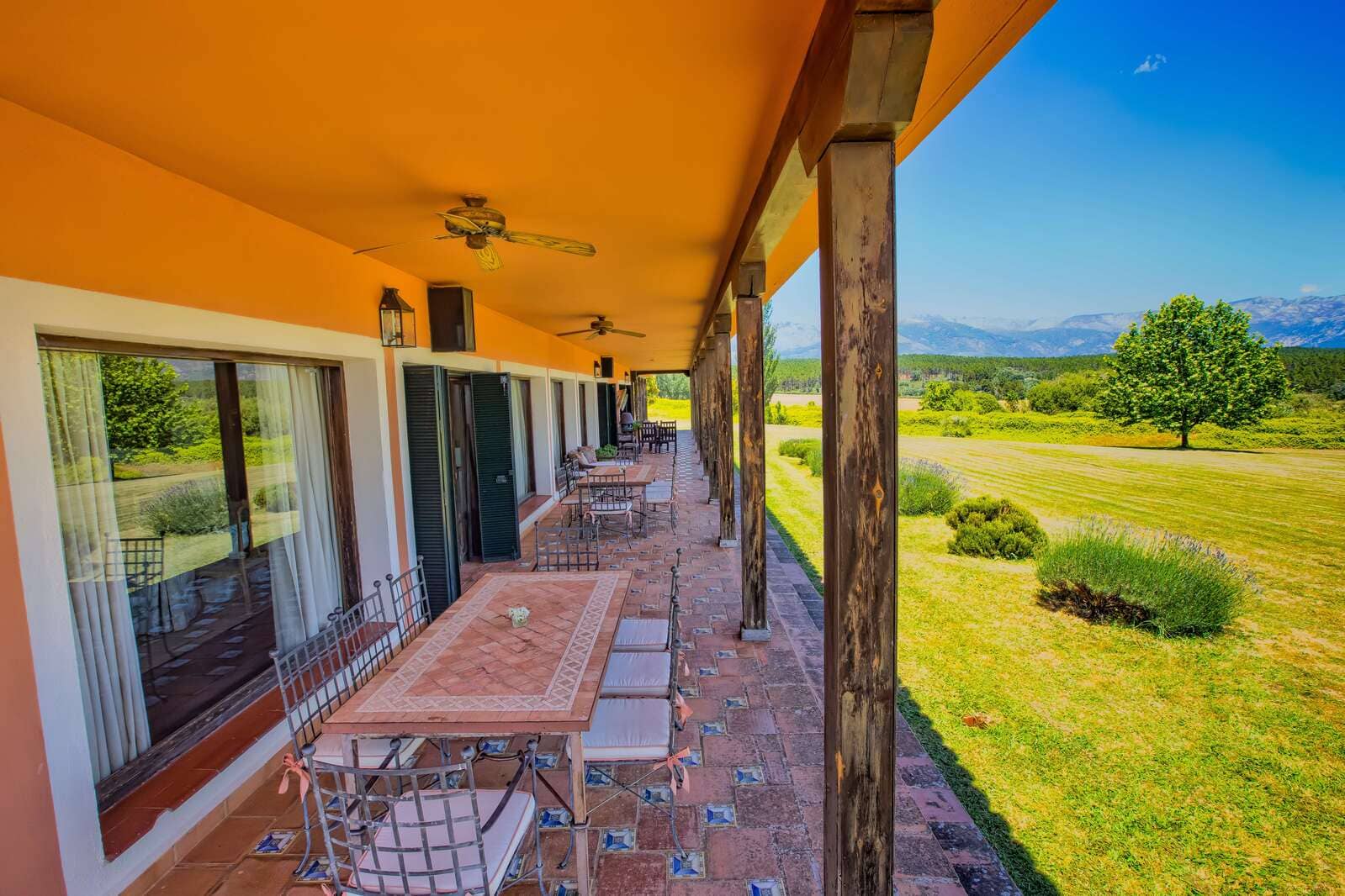 Terraza de una casa rural con techo naranja y columnas de madera, vistas al jardín y las montañas en el horizonte.