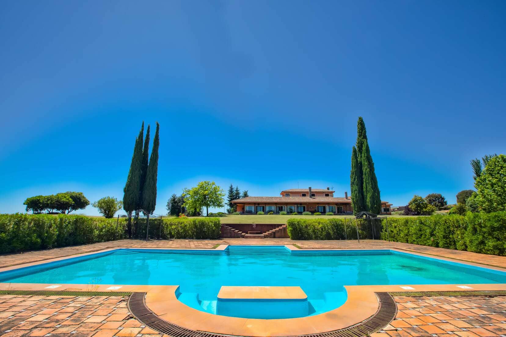 Vista de la piscina y la casa principal de una finca rural, con un cielo despejado y árboles en el fondo.