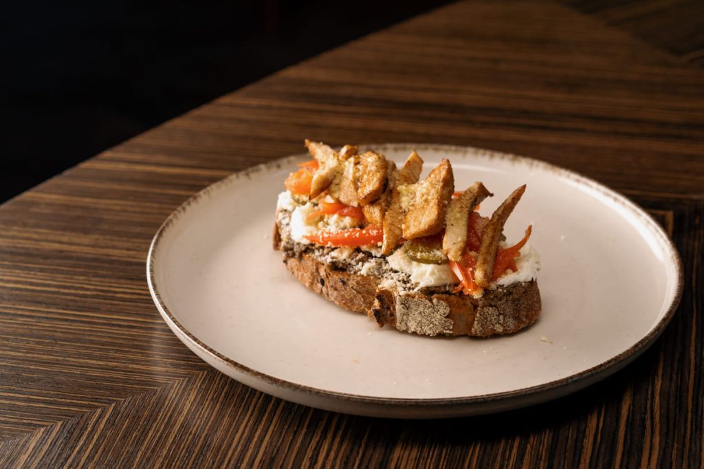 Plato gourmet de tostada con queso crema, pimientos asados, pepinos y tiras de pollo en una mesa de madera oscura.