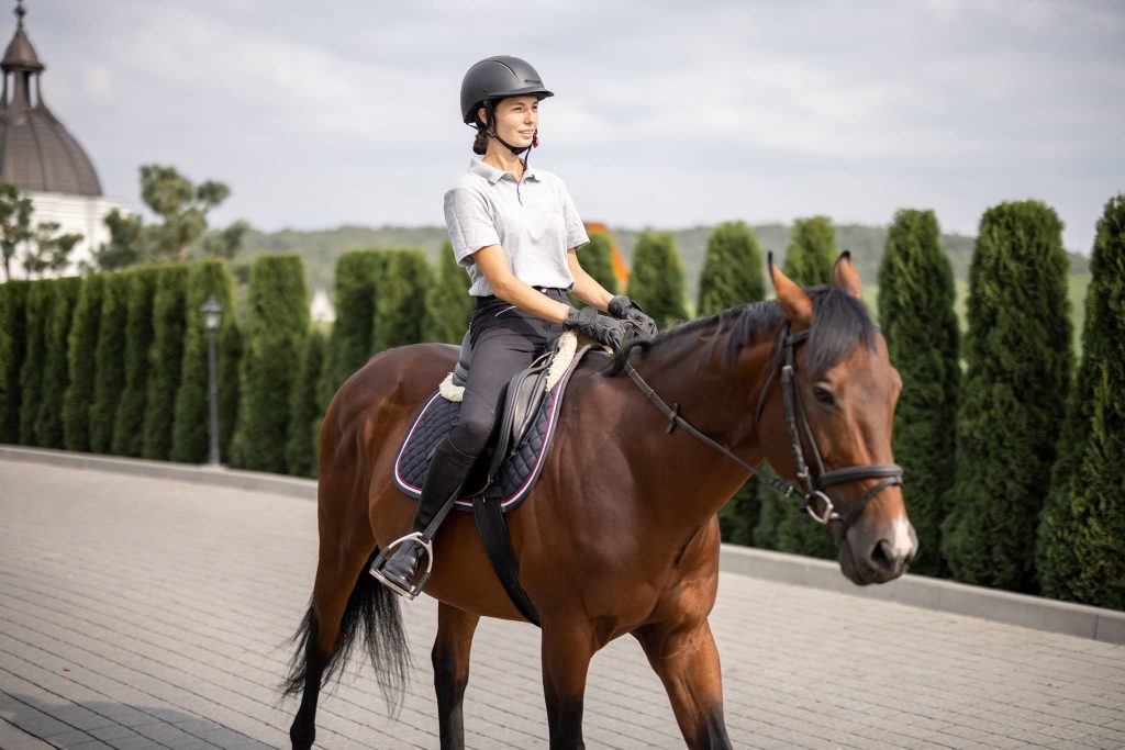 Jinete femenina montando un caballo marrón en un área pavimentada con exuberantes setos verdes y una estructura arquitectónica de fondo.