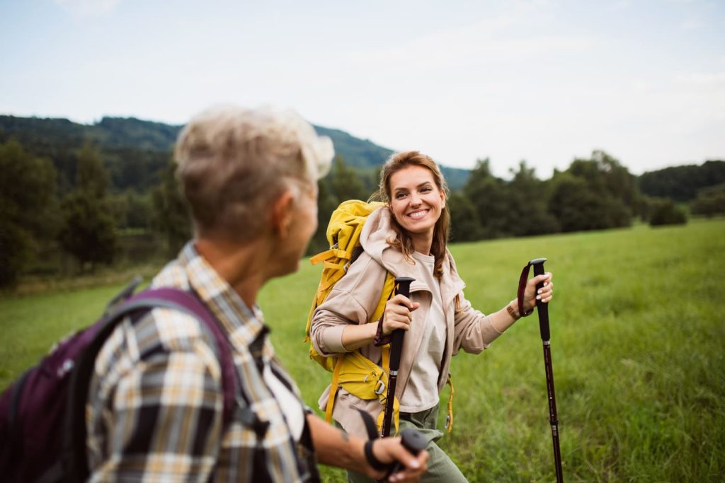 Dos excursionistas sonriendo y charlando mientras caminan por un prado verde, con montañas al fondo.