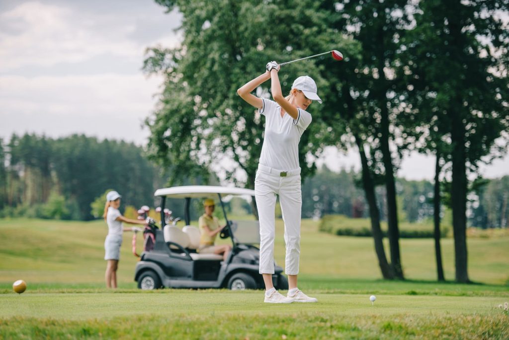 Golfista femenina en atuendo blanco completo realizando un swing en un campo de golf, con un carrito de golf y otro jugador en el fondo.